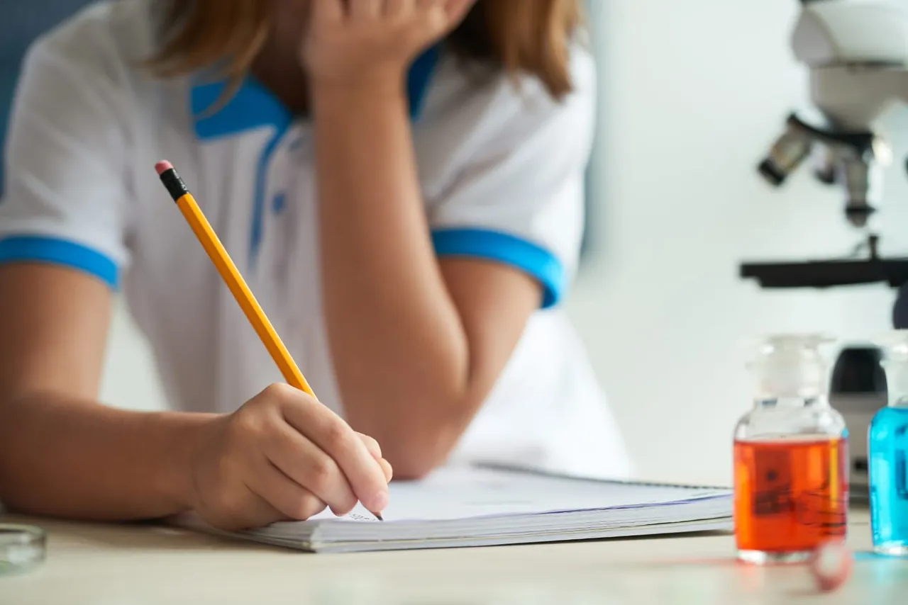 Estudiante tomando apuntes junto a un microscopio y frascos de reactivos