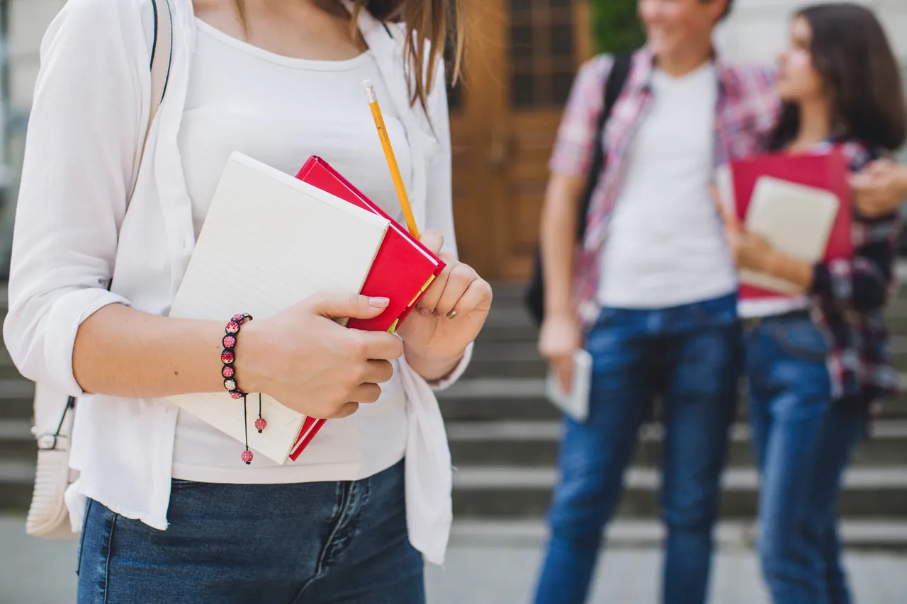Estudiante de secundaria sujetando un cuaderno y un lápiz, con compañeros al fondo en las escaleras