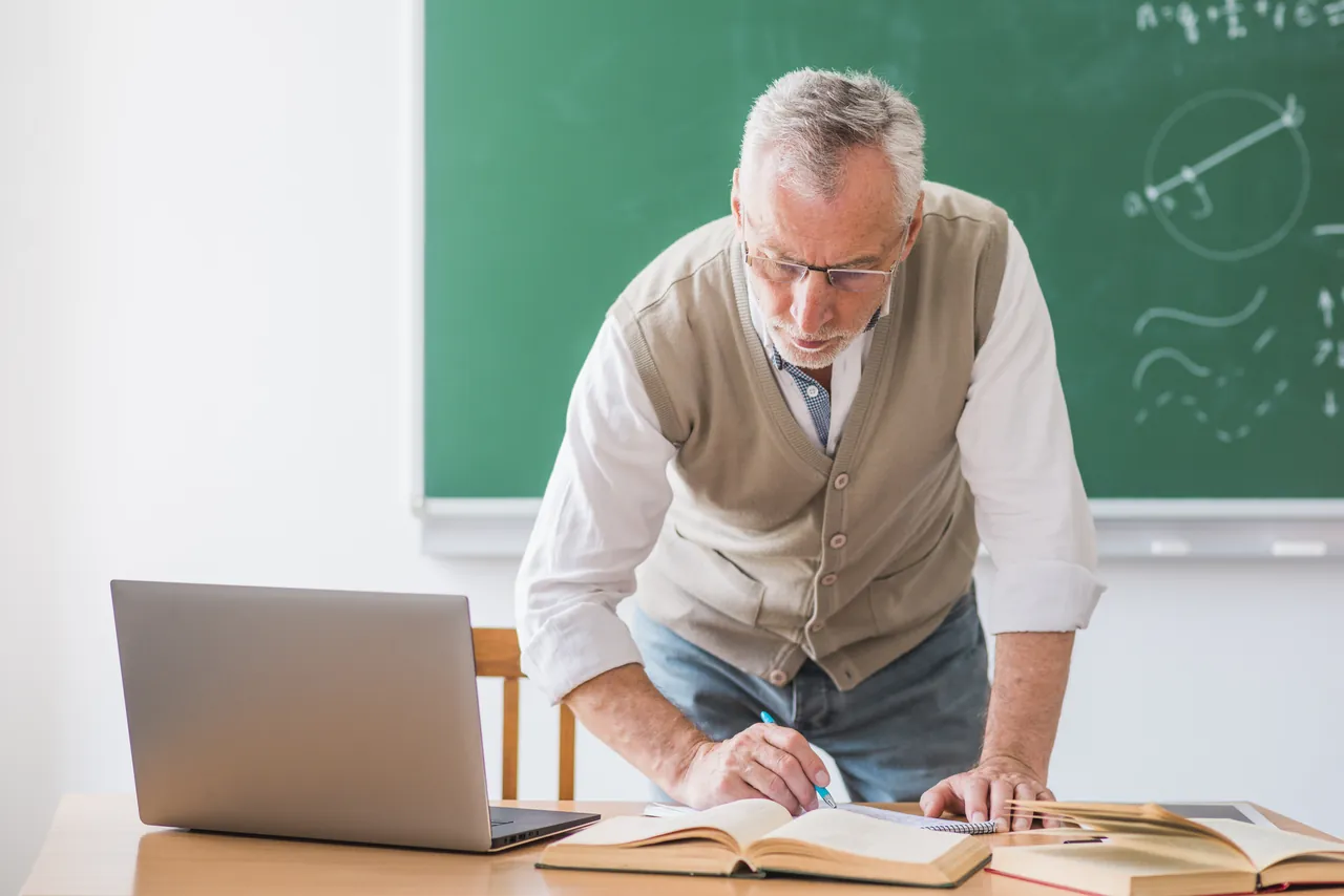 Profesor de física universitaria revisando apuntes en su mesa junto a la pizarra