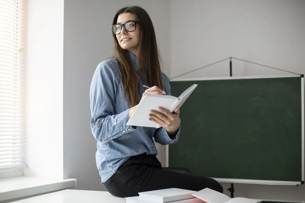 Profesora sonriente con un cuaderno sentada junto a la pizarra de un aula
